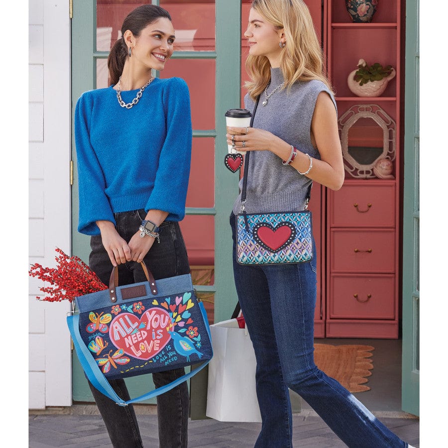 Two women stand outside a shop, smiling and talking. One holds a All You Need East West Denim Tote bag with the text "ALL YOU NEED IS LOVE" while the other holds a coffee cup and a purse with a heart design.