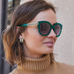 A person is wearing Intrigue Emerald Sunglasses and matching decorative hoop earrings while standing outdoors, near a textured wall and a window.