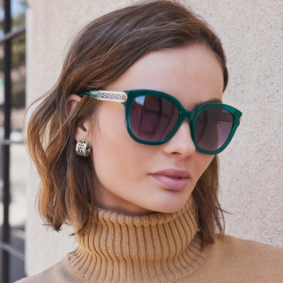 A person wearing Intrigue Emerald Sunglasses and gold earrings stands outside against a beige wall, dressed in a camel-colored turtleneck sweater.