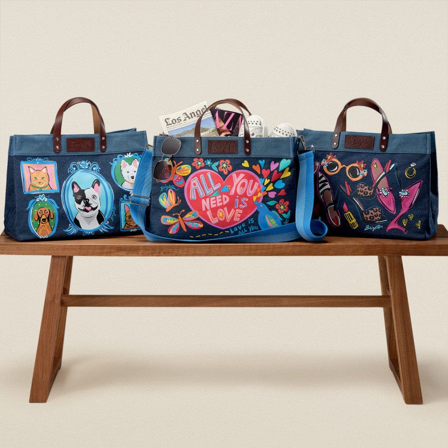 Three illustrated blue bags with leather handles sit on a wooden table. Middle bag: All You Need East West Denim Tote with "ALL YOU NEED IS LOVE" in colorful text. Surrounding items: sunglasses, and a Los Angeles newspaper.