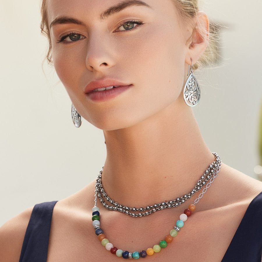 A woman wears a silver, beaded Contempo Confetti Necklace and colorful gemstone bead necklace, with silver, teardrop-shaped earrings, all set against a soft, neutral background with part of a plant visible.