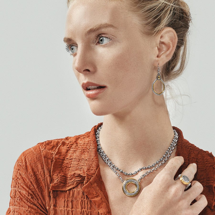 A woman wearing Venezia Teardrop French Wire Earrings, a textured orange top showcases a necklace with a large circular pendant and a ring. She is looking to the side against a neutral background.